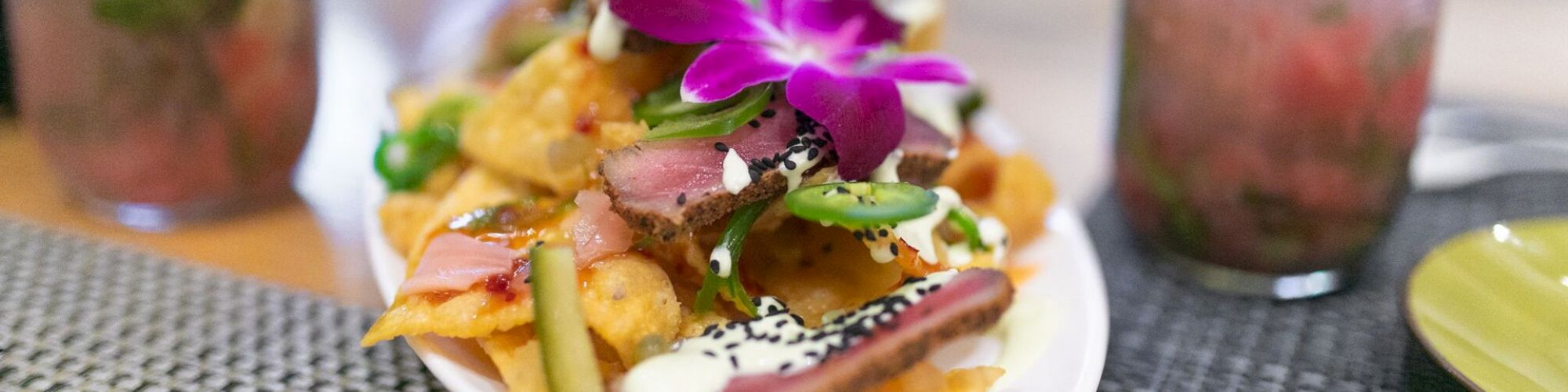 A plate of nachos topped with sliced seared tuna, green peppers, and a purple flower, with drinks in jars on the side.