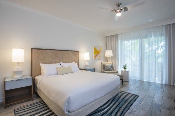 A modern bedroom with a large bed, two bedside tables with lamps, an armchair, a rug, and large windows with curtains letting in natural light.