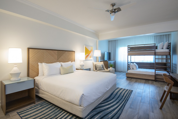 A spacious, modern bedroom features a king-size bed, a set of bunk beds, a desk area, and a seating area, all with stylish decor and ample lighting.