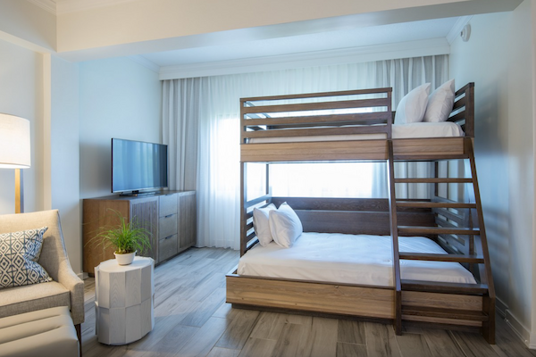 The image shows a modern room with a bunk bed, TV on a wooden cabinet, a white side table with a plant, and an armchair with a pillow.