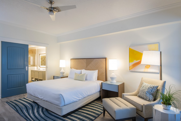 A modern bedroom featuring a neatly made bed, side tables with lamps, an armchair, artwork, and a view into an adjoining bathroom.