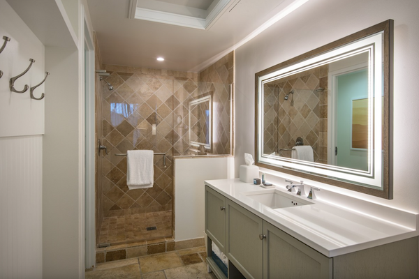 This image shows a modern bathroom with a large mirror, a double sink vanity, hooks on the wall, and a walk-in shower with tiled walls and a towel hanging.