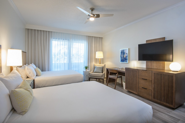 The image shows a hotel room with two beds, a flat-screen TV, a desk with a chair, and a window covered by light curtains.