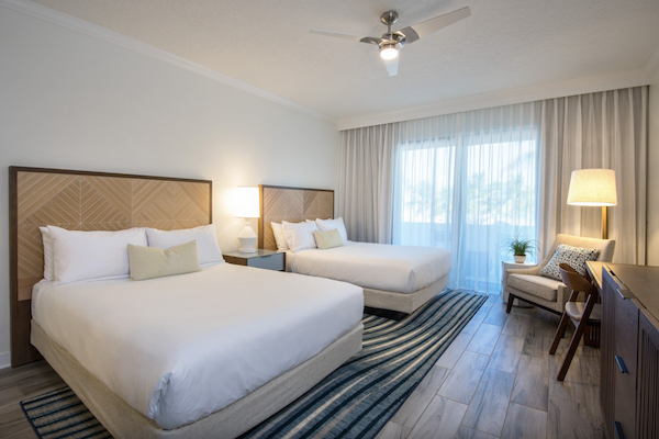 A modern hotel room features two double beds, a ceiling fan, a desk, chairs, lamps, and large windows with sheer curtains for natural light.