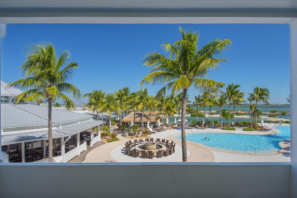 A resort scene with palm trees, a pool, seating area, cabanas, and ocean view under a clear blue sky.