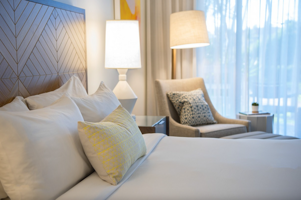 A cozy, well-lit bedroom with a neatly made bed, two lamps, an armchair, a side table, and sheer curtains allowing natural light.