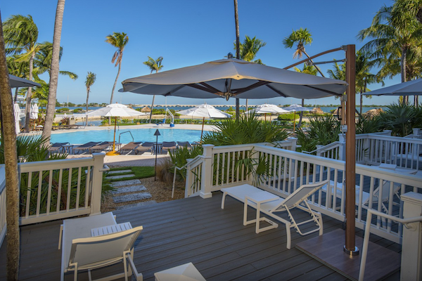 The image showcases a tropical poolside area with lounge chairs, a deck, and large umbrellas, surrounded by palm trees on a sunny day.