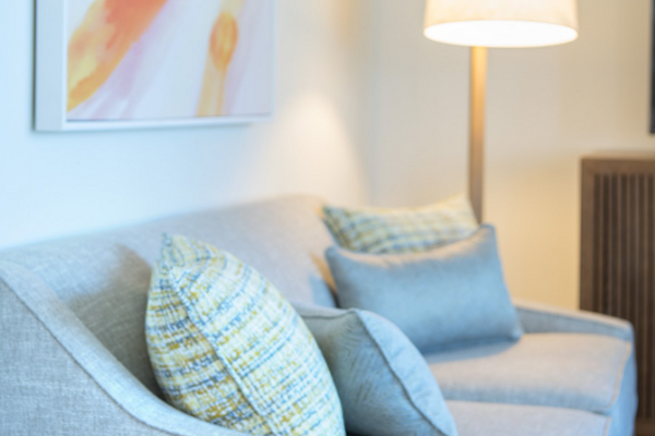 A cozy living room with a grey sofa, colorful pillows, a small side table with a plant, a standing lamp, and abstract wall art in the corner.