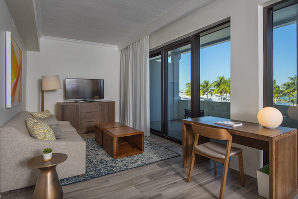 The image shows a modern living room with a sofa, TV, desk, chair, coffee table, lamp, and large windows with views of palm trees outside.