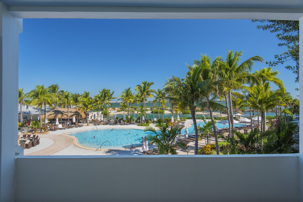 The image shows a resort view featuring a swimming pool, palm trees, lounge chairs, and clear blue skies, framed by a window-like opening.