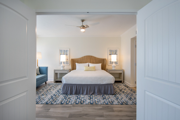 A bright bedroom with a large bed, blue sofa, nightstands, lamps, and a ceiling fan, viewed through double doors.