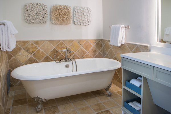 The image shows a bathroom with a freestanding bathtub, wall decor, towels on racks, tiled walls and floor, and a vanity with storage shelves.