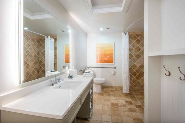 A modern bathroom with a double sink, large mirror, tiled shower area, toilet, floor tiles, hooks, and an orange-themed artwork on the wall.