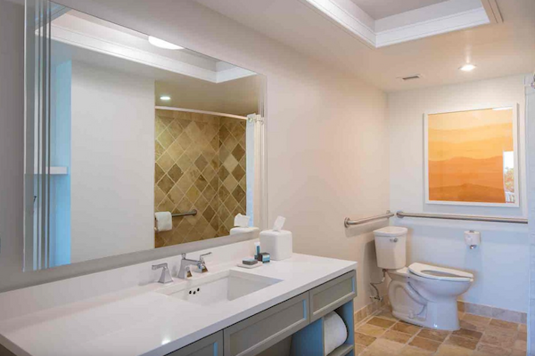 A modern bathroom with a large mirror, double sink, shower with tiled walls, and a toilet next to a framed artwork on the wall.