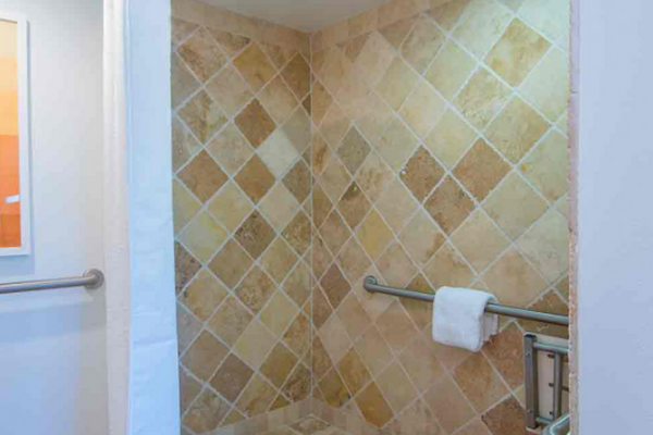 The image shows a walk-in shower with beige wall tiles, a shower curtain, a grab bar, and a towel rack holding a white towel.