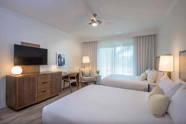 This is a modern hotel room with two beds, a TV, a desk, lamps, and a chair, featuring light decor and a ceiling fan in the center.