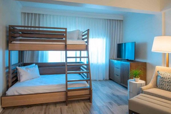 The image shows a cozy room with a wooden bunk bed, a TV on a stand, a chair with a pillow, a plant on a side table, and a window with curtains.