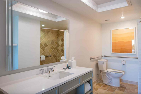 A modern bathroom with a large mirror, sink, toilet, walk-in shower, and beige tiles, with stylish lighting and art on the wall.