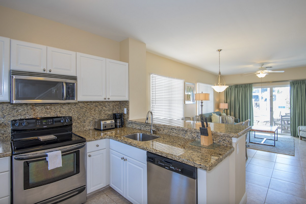 A modern kitchen with granite countertops, white cabinets, and stainless steel appliances, opening into a living room with large windows.