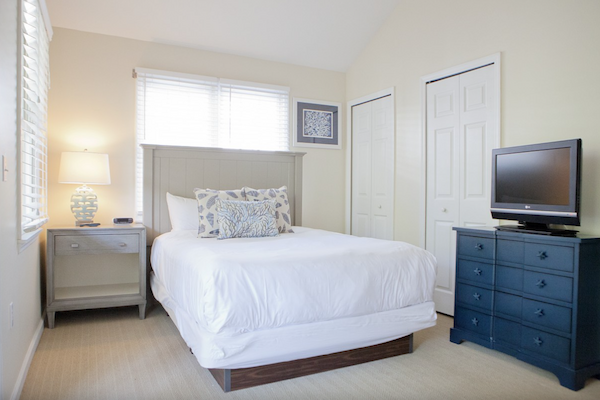 This image shows a bedroom with a bed, bedside table with a lamp, chest of drawers with a TV, and two closets. The room has a bright and clean look.