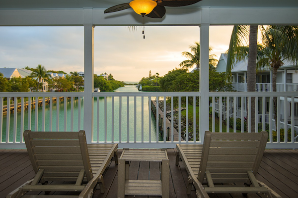 A covered porch features two lounge chairs facing a serene waterway with lush greenery and houses, under a ceiling fan, at sunset.