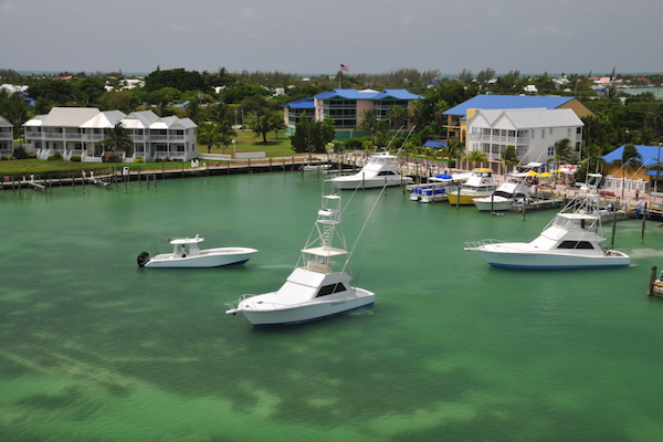 The image shows a marina with several boats docked on clear turquoise water, surrounded by waterfront homes and lush greenery.