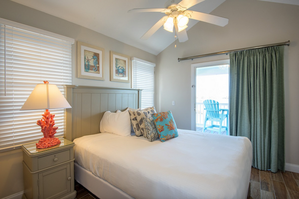 A cozy bedroom with a bed, bedside table, coral lamp, pictures on the wall, ceiling fan, and a sliding door leading to a balcony with chairs.