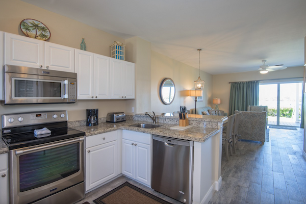 A modern kitchen and dining area with white cabinets, stainless steel appliances, granite countertops, and a view of a living room leading to a balcony.