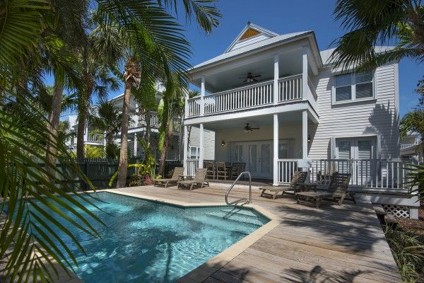 A two-story house surrounded by palm trees features a spacious patio with lounge chairs and a swimming pool in the foreground, under clear skies.