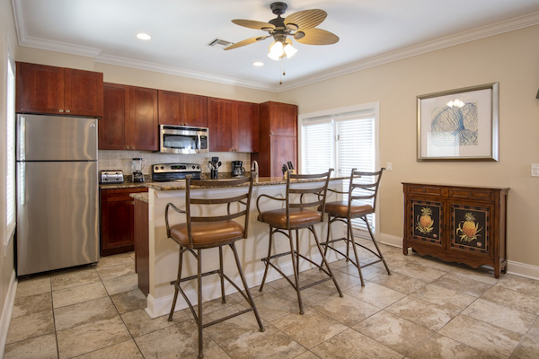 A kitchen with wooden cabinets, stainless steel appliances, bar stools at an island, tiled floor, ceiling fan, and a side cabinet with a framed picture.