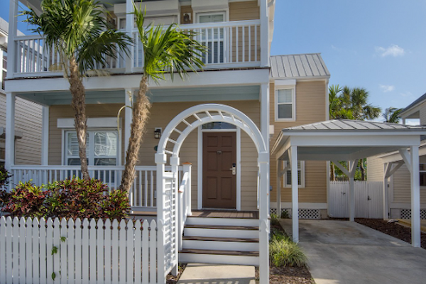 A charming two-story house with a white picket fence, front porch, balcony, and driveway under clear blue skies.