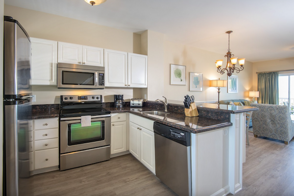 A modern kitchen with stainless steel appliances, white cabinets, a countertop with a knife set, and a view into the living area with a chandelier.