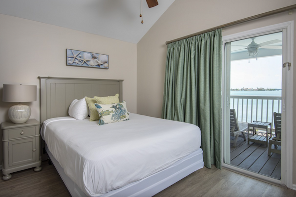 A cozy bedroom with a neatly made bed, nightstand, lamp, and a sliding glass door leading to a balcony overlooking a scenic body of water.