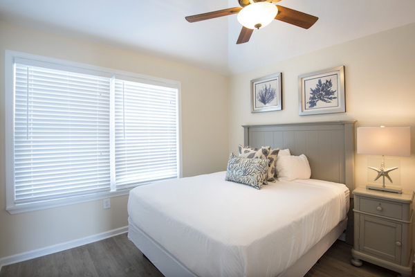 The image shows a neatly arranged bedroom with a ceiling fan, a bed with pillows, two framed pictures above the headboard, a nightstand, and a window.