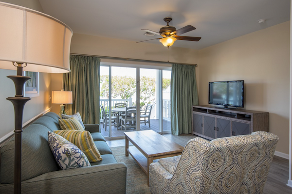 A cozy living room with a sofa, armchair, lamp, TV, and patio doors leading to a balcony with outdoor seating.