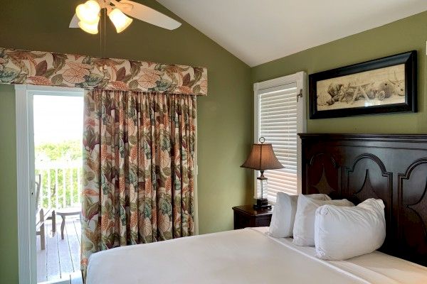 A bedroom with a bed, floral curtains, a ceiling fan, a lamp, a framed picture, and a partially open door leading to a balcony with outdoor furniture.