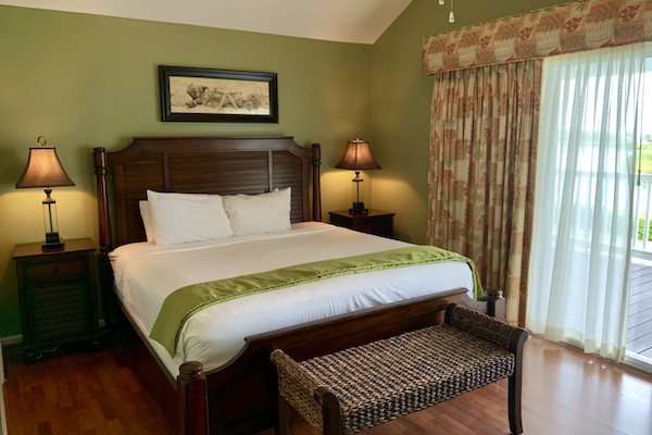 A cozy bedroom with a large bed, two side tables with lamps, a wicker bench at the foot of the bed, and a large window with curtains.