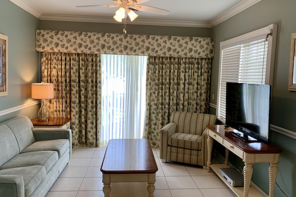 A cozy living room with a green sofa, armchair, coffee table, lamp, TV, and patterned curtains covering sliding doors to a balcony.