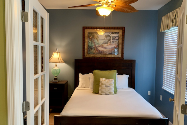 A cozy bedroom features a bed with green pillows, a side table with a lamp, a ceiling fan, a painting above the bed, and blue walls ending the sentence.