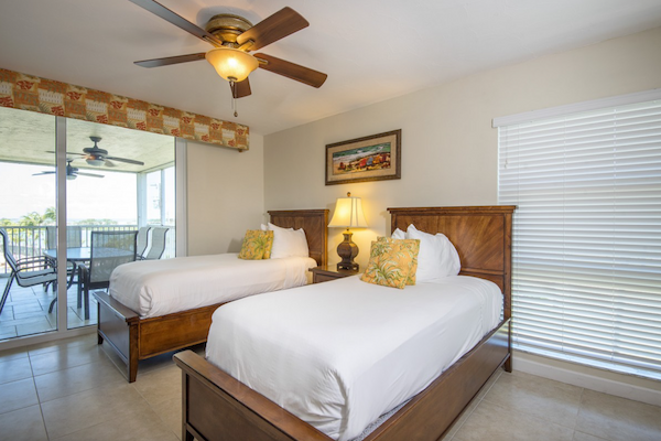 A bright bedroom with two single beds, a ceiling fan, and a lamp between the beds, with a sliding door leading to an outdoor balcony.