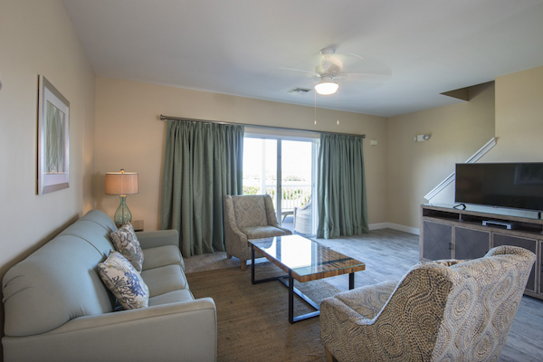 A living room with a sofa, two chairs, coffee table, TV, and large window with curtains. Light walls and ceiling fan complete the space.