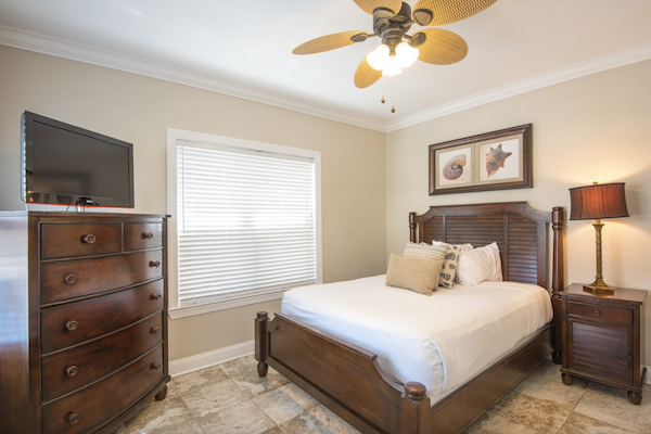 A bedroom with a wooden bed, dresser, nightstand, lamp, ceiling fan, and wall art, plus a window and TV.