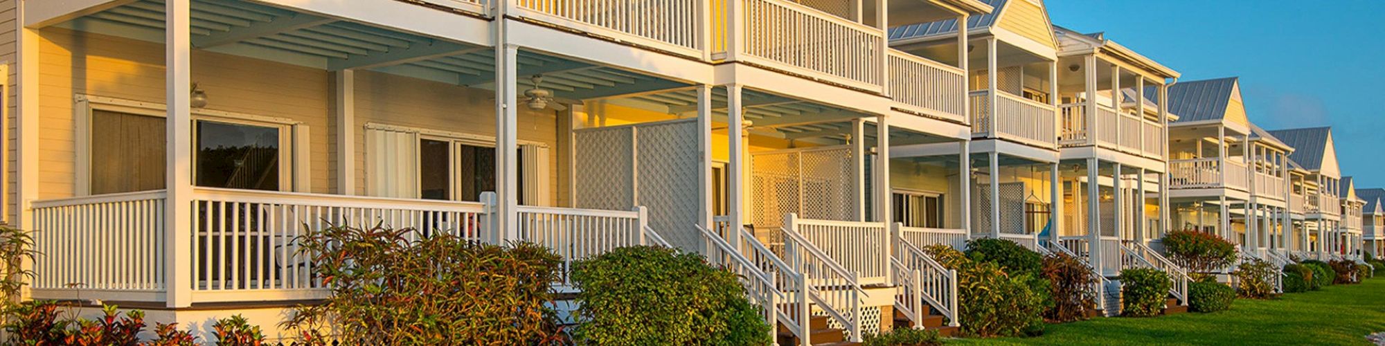 The image depicts a row of beige, two-story residential buildings with white balconies, railings, and well-maintained lawns in the foreground.