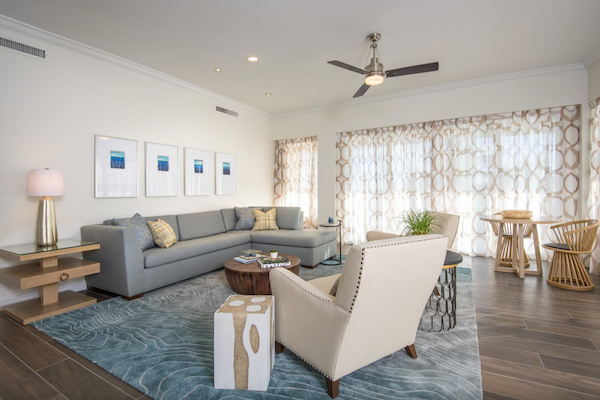 A modern living room with a grey sectional sofa, patterned armchair, wooden furniture, blue rug, wall art, and large windows with geometric curtains.