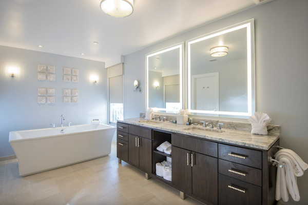 The image shows a modern bathroom with a freestanding tub, double vanity with illuminated mirrors, and soft lighting, creating a luxurious feel.