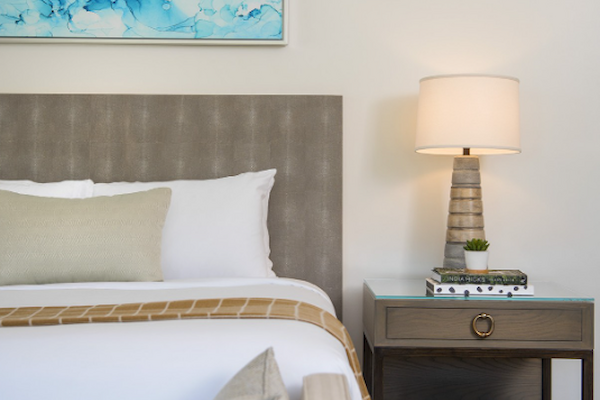 A modern bedroom showcases a bed with pillows, an abstract blue painting, and a lamp on a nightstand with books and decor.