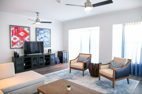 A modern living room with two armchairs, a TV, abstract wall art, ceiling fans, and a coffee table on a rug, all in a bright setting.