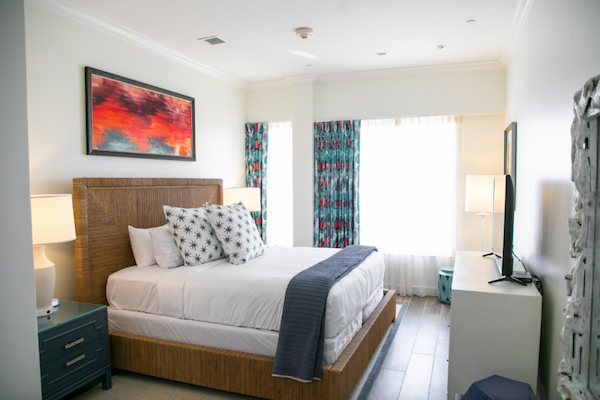 A bright, modern bedroom with a cozy bed, vibrant artwork, two bedside tables with lamps, colorful curtains, and a TV on a white dresser.