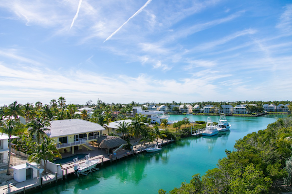 A tropical waterfront scene with houses, palm trees, boats, and clear water, set against a bright blue sky with wispy clouds.