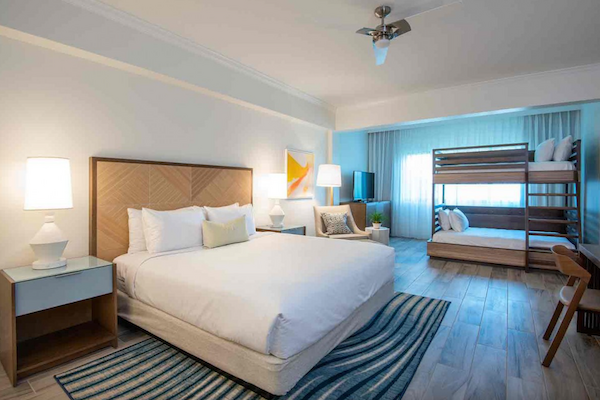 A modern hotel room with a large bed, nightstands, bunk beds, a sitting area, a desk, lamps, and a rug on wooden flooring, in white and blue tones.